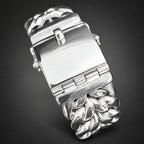 A close-up of a silver-toned metal watch band being held in a hand, showing the clasp and textured leaf-like links.