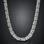 A silver-toned interlocking chain necklace with a braided or woven link design, shown draped against a dark gray gradient background.