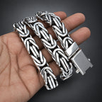 A silver-toned braided chain link bracelet with a polished rectangular clasp, displayed on a dark gray gradient background.