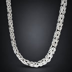A silver-toned braided chain necklace with a polished finish, arranged in a U-shape against a dark gray gradient background.