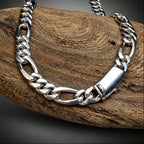 A silver-toned chunky curb chain necklace with a polished rectangular clasp, draped over a textured brown wooden log against a dark gray gradient background.