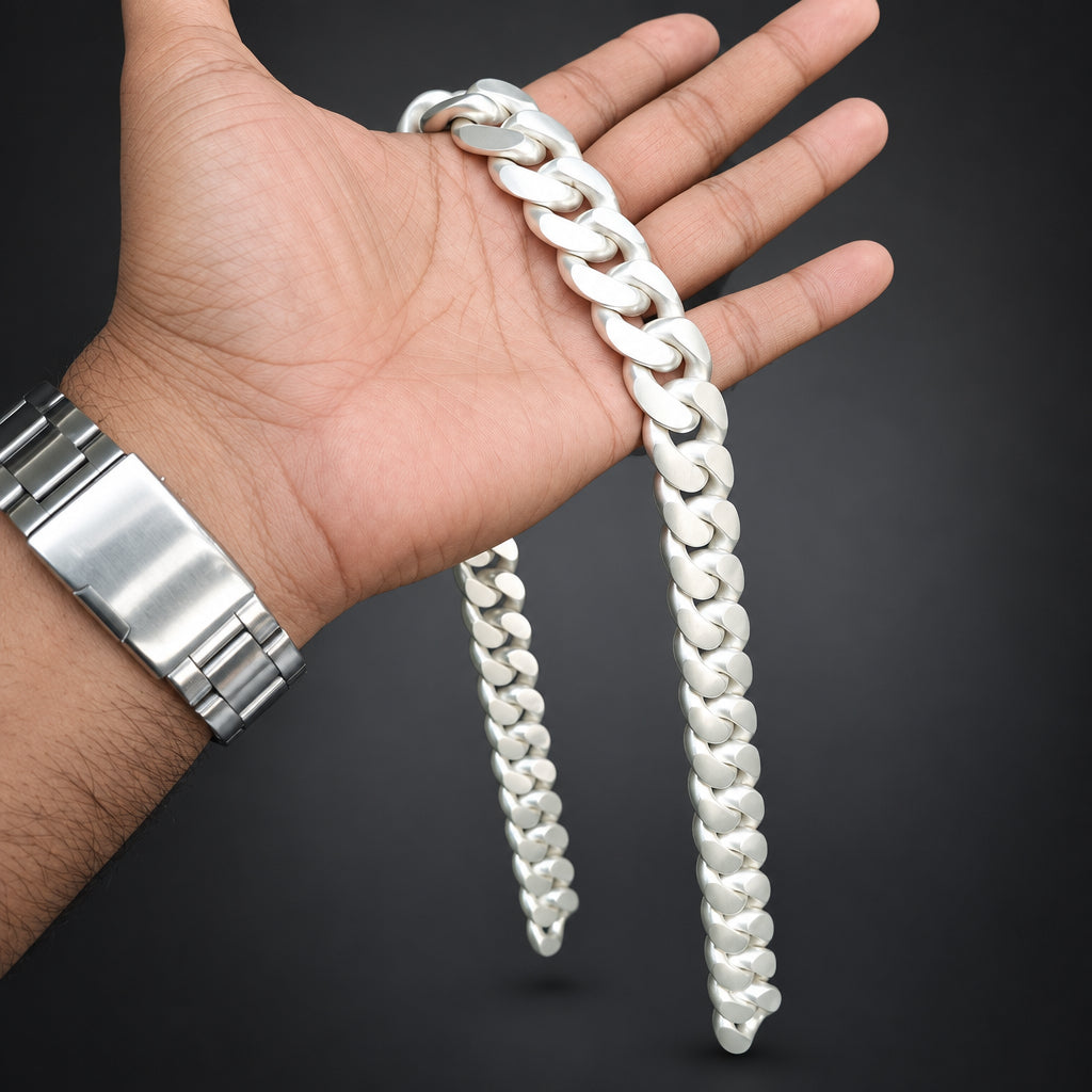 A silver Cuban link chain necklace draped over a person's hand, with a matching silver metal watch visible on the wrist, set against a dark gray background.