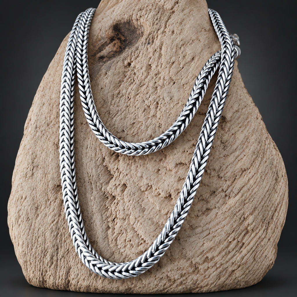 A silver braided chain necklace draped over a textured beige stone display against a dark background.