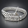 A silver-toned Cuban link chain bracelet with an ornate engraved clasp, displayed on a textured beige surface against a dark background.