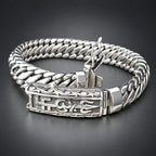 A silver-toned Cuban link chain bracelet with an ornate engraved clasp, displayed on a textured beige surface against a dark background.