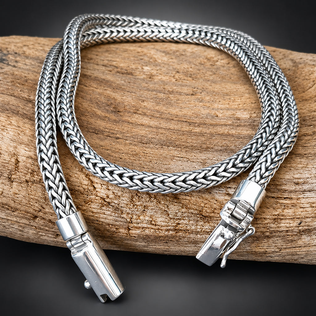 A silver braided snake chain necklace with a lobster clasp, displayed on a piece of weathered driftwood against a dark background.
