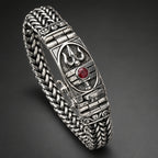 A close-up of a silver-toned metal bracelet worn on a person's wrist. The bracelet features an ornate shield-like clasp with a small round red gemstone in the center and a braided mesh band.