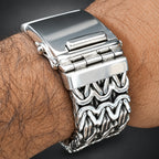 A close-up of a silver-toned metal link watch band worn on a person's wrist, showing the clasp and textured wavy links against dark skin and a black background.