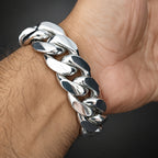 A close-up of a shiny silver Cuban link chain bracelet worn on a person's wrist, with the hand and arm visible against a dark background.