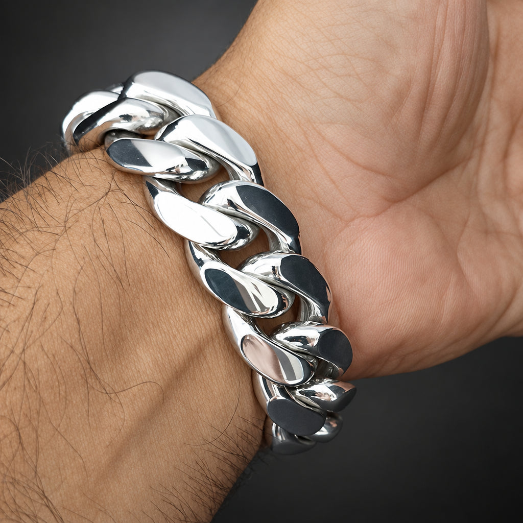 A close-up of a shiny silver Cuban link chain bracelet worn on a person's wrist, with the hand and arm visible against a dark background.