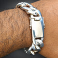A close-up of a chunky silver Cuban link chain bracelet worn on a hairy wrist, featuring a polished rectangular clasp.