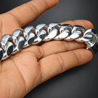 A close-up of a hand holding a chunky, highly polished silver Cuban link chain bracelet against a dark gray background.