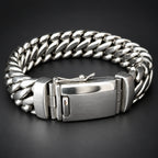 A silver stainless steel Cuban link bracelet with a brushed metal clasp, displayed on a dark gray background.