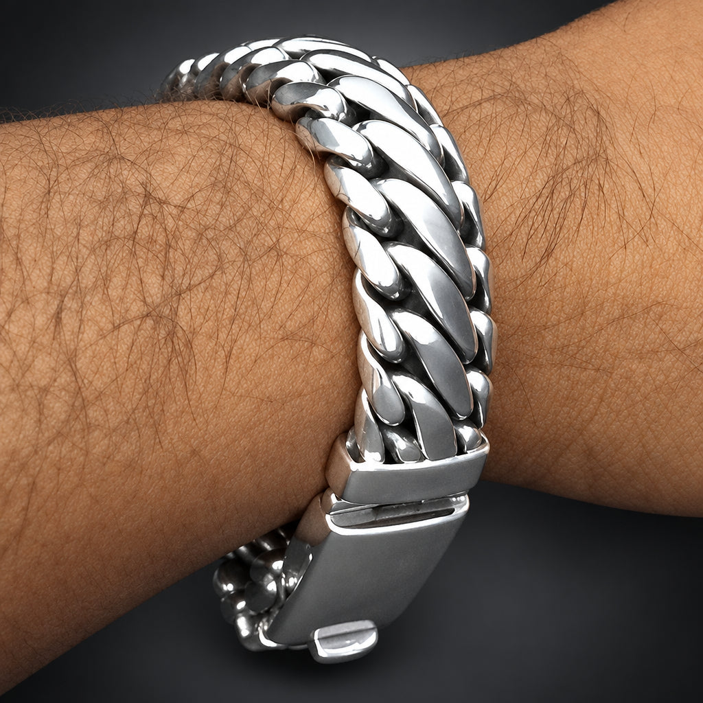 A close-up of a silver-toned Cuban link chain bracelet worn on a person's wrist, showing the textured links and clasp against dark skin and a black background.