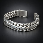 A silver-toned Cuban link chain bracelet with a polished finish and a rectangular clasp, displayed on a dark textured surface.