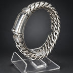 A silver-toned metal Cuban link bracelet displayed on a clear acrylic stand against a dark gray background.