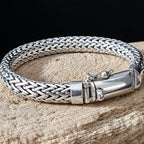 A silver braided chain bracelet with a polished clasp, resting on a textured beige stone surface against a black background.