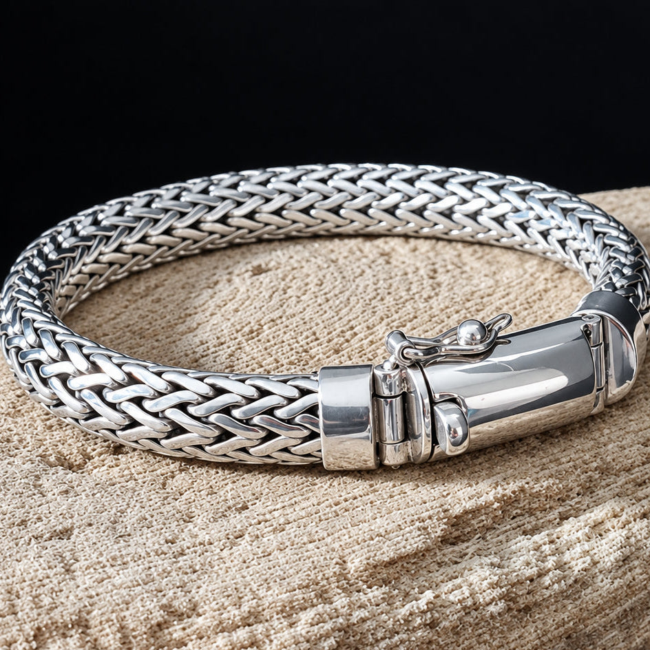 A silver braided chain bracelet with a polished clasp, resting on a textured beige stone surface against a black background.