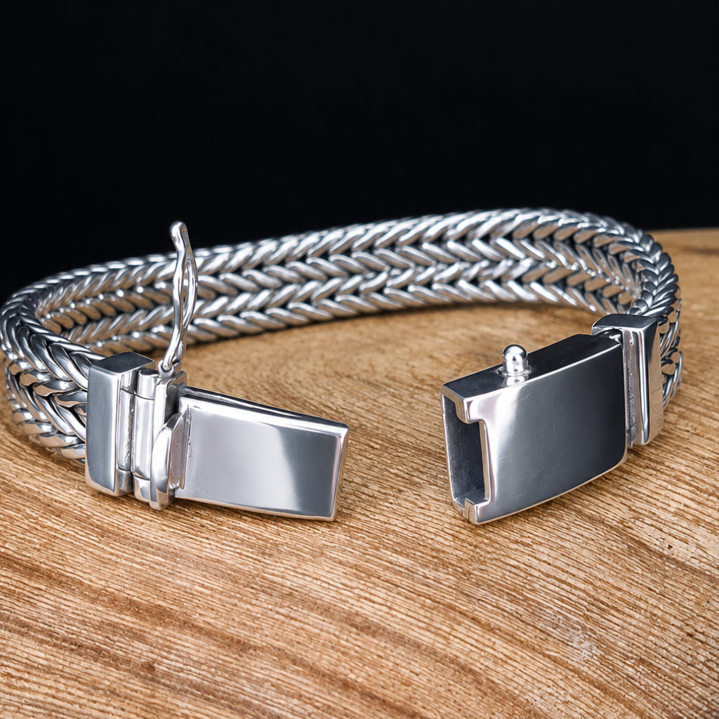 A silver-toned braided metal cuff bracelet with two rectangular slide clasp ends, displayed on a round wooden slice against a black background.