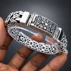A close-up of a hand holding a silver-toned bangle bracelet with an ornate engraved clasp and patterned chain links, shown against a dark gray gradient background