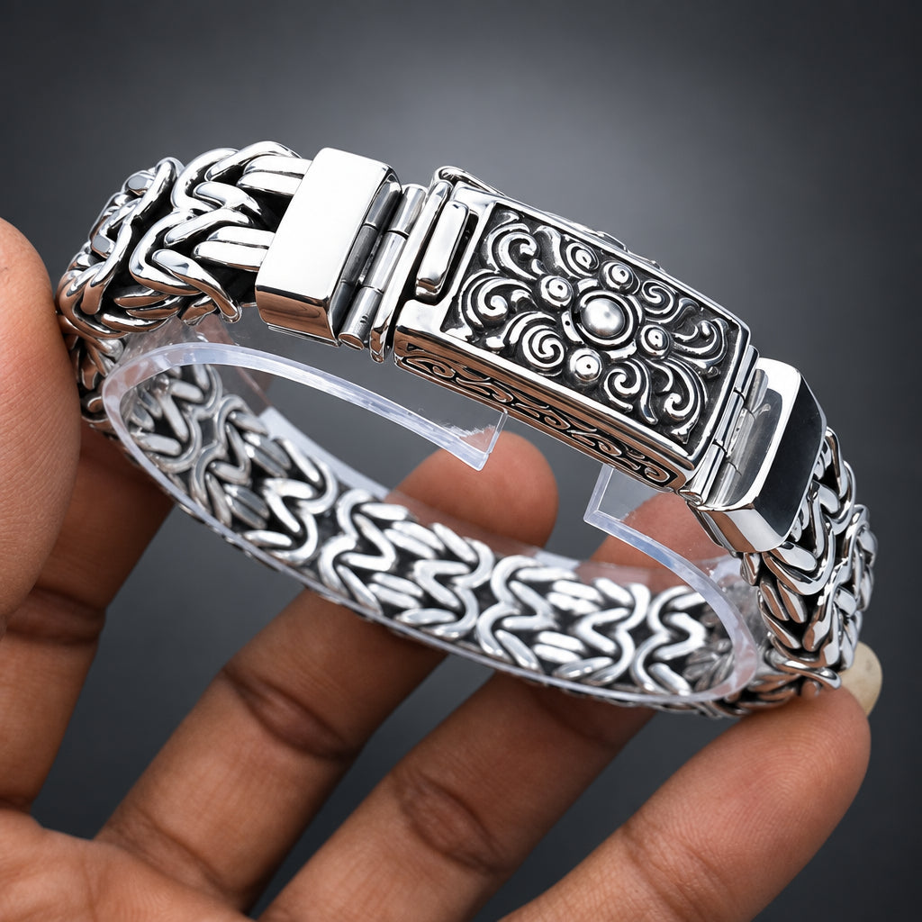 A close-up of a hand holding a silver-toned bangle bracelet with an ornate engraved clasp and patterned chain links, shown against a dark gray gradient background
