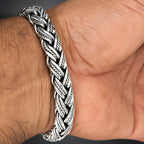 A close-up of a thick braided silver-toned metal bracelet worn on a person's wrist, showing the textured woven pattern against skin.
