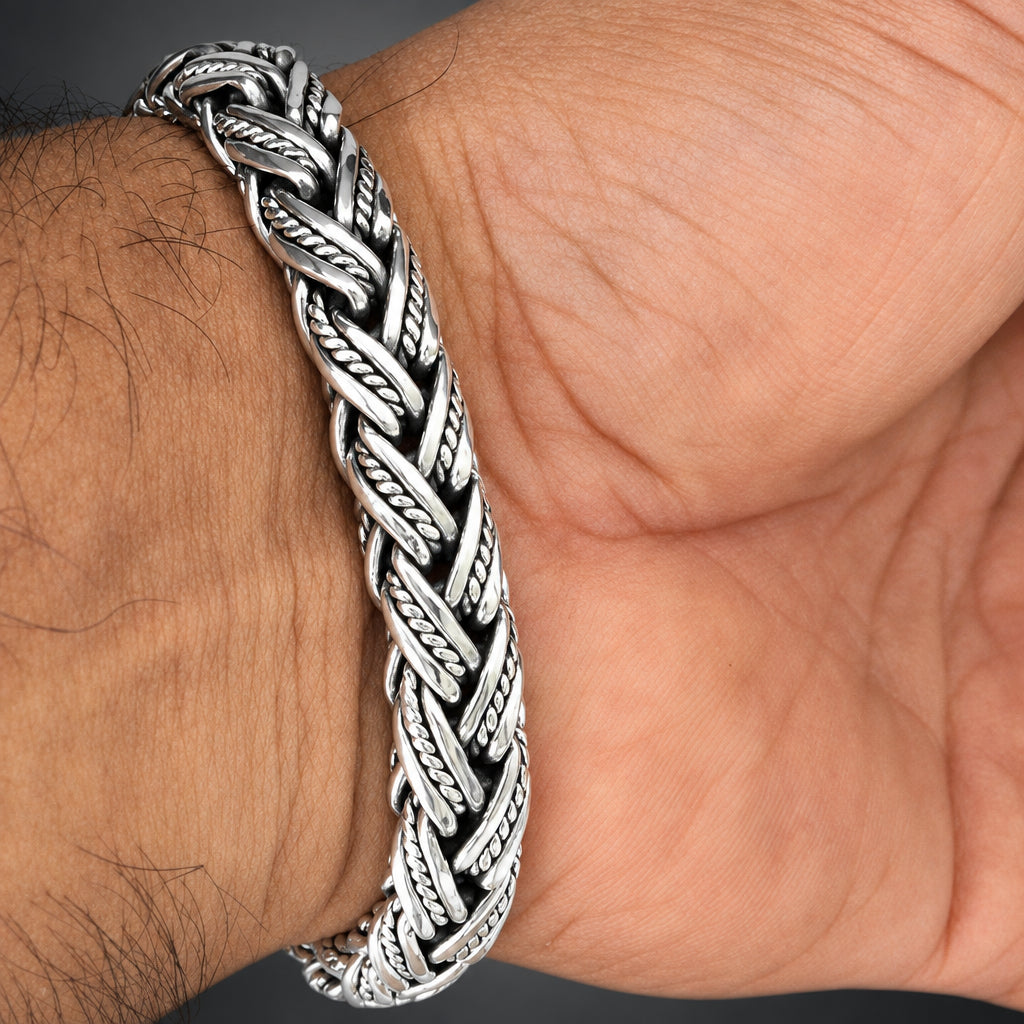 A close-up of a thick braided silver-toned metal bracelet worn on a person's wrist, showing the textured woven pattern against skin.