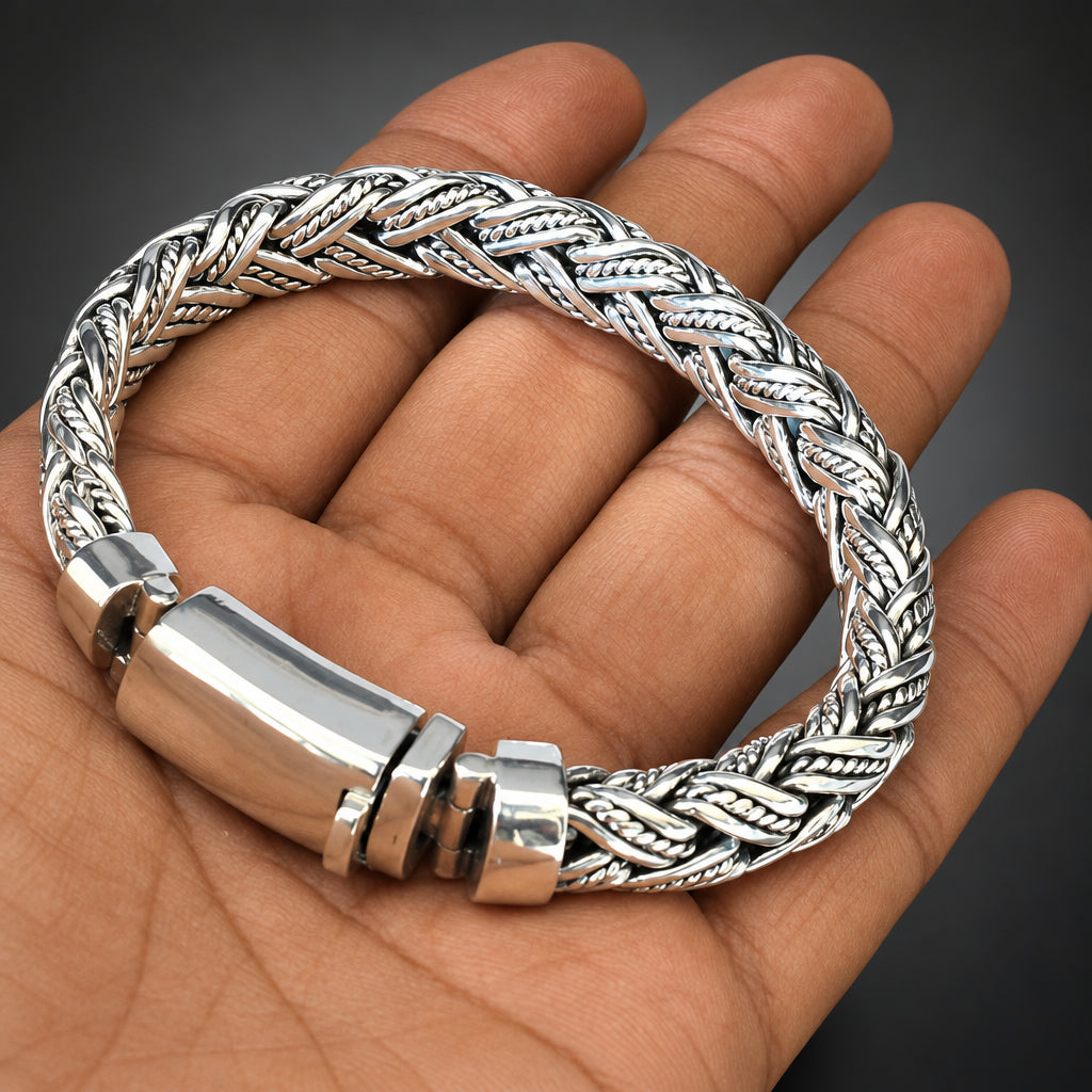 A silver braided chain bracelet with a polished cylindrical clasp, shown resting on a person's hand.