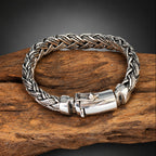 A braided silver-toned metal bracelet with a polished clasp, displayed on a piece of rustic wood against a dark gray gradient background.
