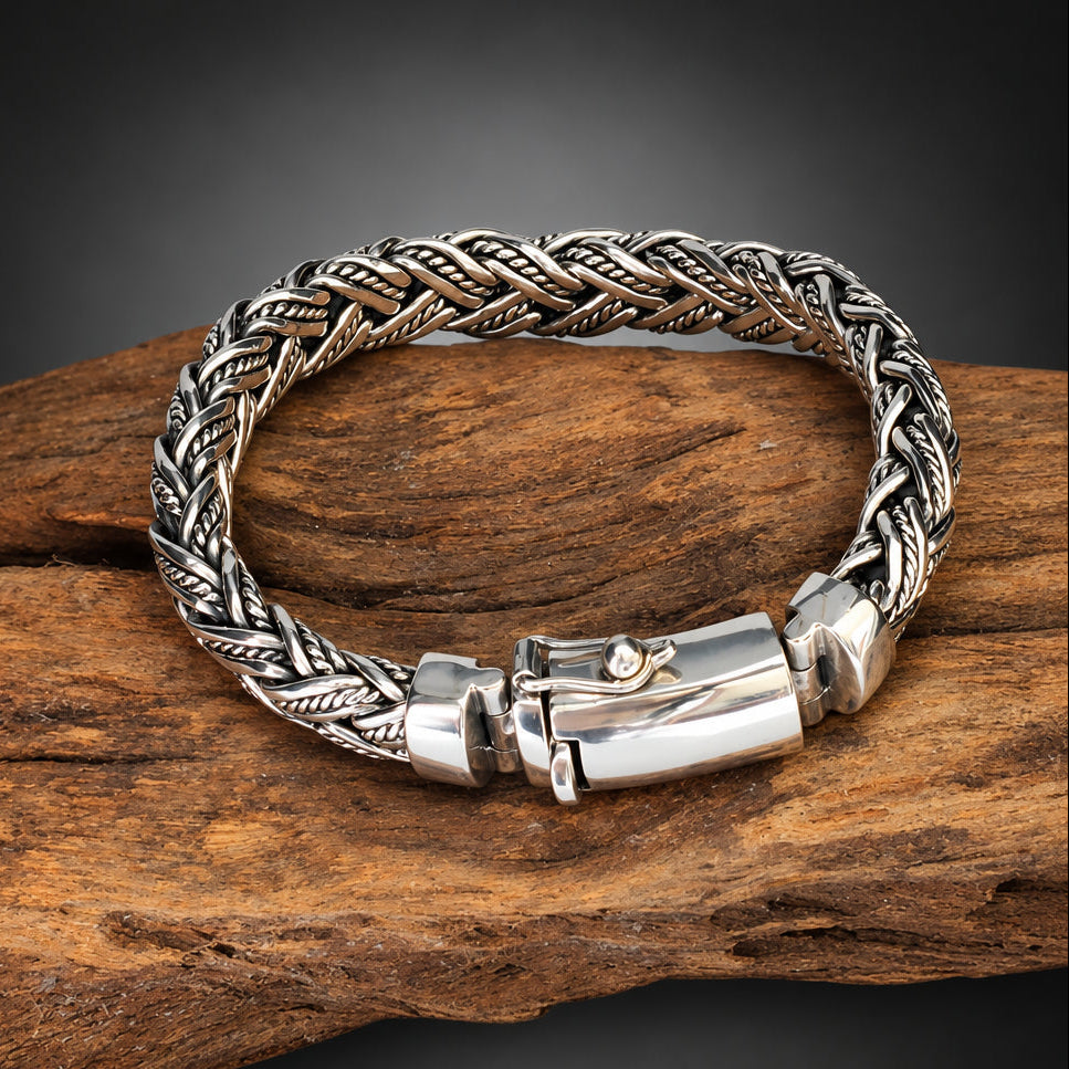 A braided silver-toned metal bracelet with a polished clasp, displayed on a piece of rustic wood against a dark gray gradient background.