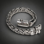 A silver-toned braided chain bracelet with a polished rectangular clasp, displayed on a neutral light gray background.