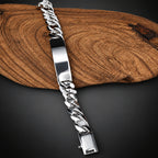 A silver Cuban link chain ID bracelet with a polished rectangular nameplate, displayed diagonally on a rustic wood slice over a white background.