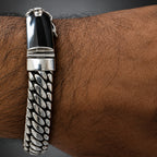 A men's silver-toned chain bracelet with a polished rectangular black inlay clasp, shown worn on a man's wrist against a dark gray gradient background.
