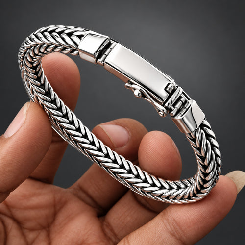 A silver-toned braided bracelet with a polished clasp, held in a person's hand against a light gray background.