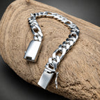 A silver Cuban link chain bracelet with polished rectangular clasps, displayed on a textured piece of driftwood against a dark gray background.