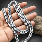 A close-up of a silver-toned curb chain necklace draped over a person's hand, with a piece of driftwood in the background.