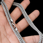 A close-up of a silver braided chain necklace draped over a person's hand against a black background.