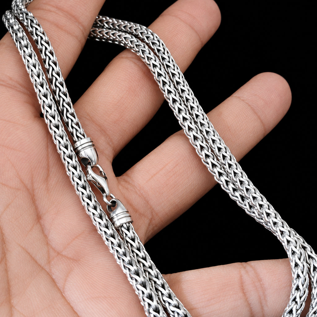 A close-up of a silver braided chain necklace draped over a person's hand against a black background.