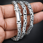 A close-up of a hand holding three braided silver-toned metal bracelets against a dark background.