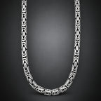 A close-up studio shot of a silver-toned chain necklace with repeating geometric interlocking links, arranged in a U-shape against a dark gray gradient background.