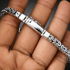 A silver-toned braided chain bracelet with a polished rectangular clasp, shown worn on a person's hand.