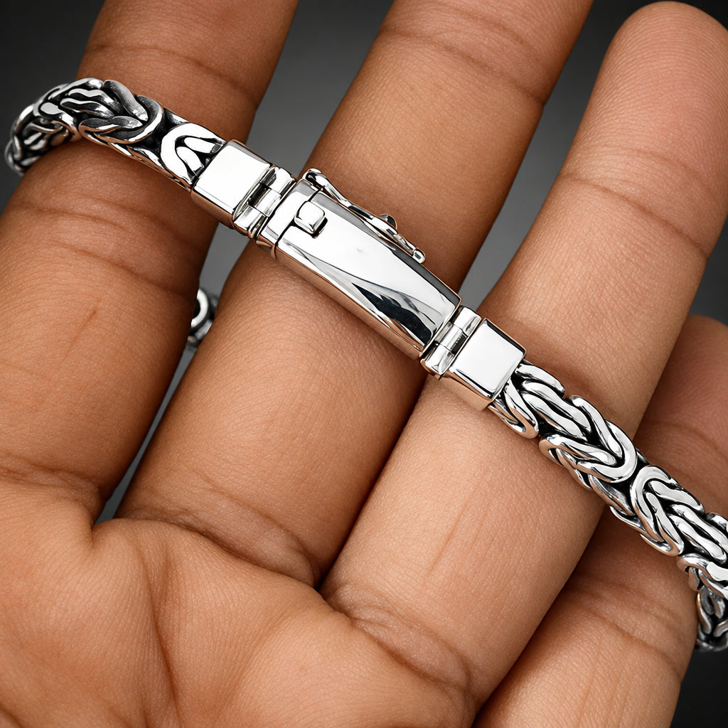 A silver-toned braided chain bracelet with a polished rectangular clasp, shown worn on a person's hand.