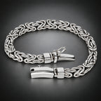 A silver-toned braided chain bracelet with a polished rectangular clasp, displayed on a dark gray gradient background.