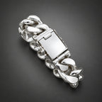 A close-up of a hand holding a chunky silver chain bracelet with a polished rectangular clasp against a neutral gray background.