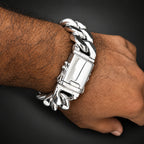A close-up of a person's wrist and hand wearing a chunky silver Cuban link bracelet with a rectangular clasp, set against a solid black background.