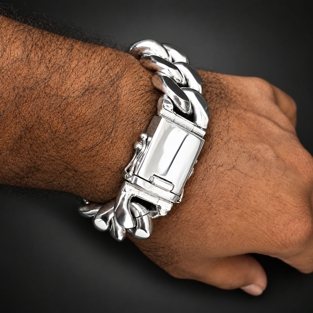 A close-up of a person's wrist and hand wearing a chunky silver Cuban link bracelet with a rectangular clasp, set against a solid black background.
