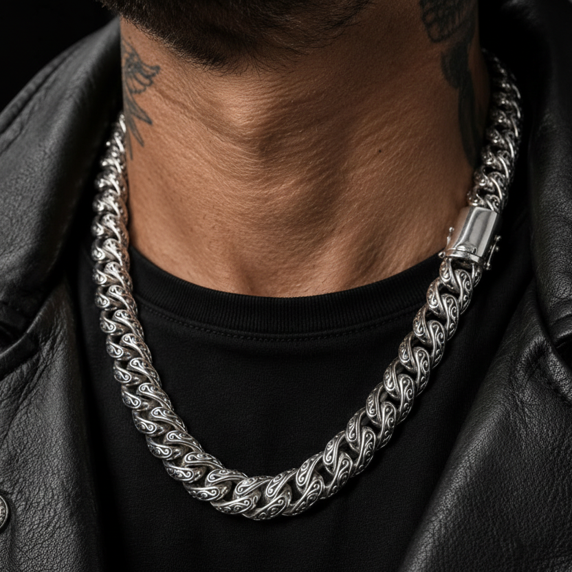 A close-up of a man's neck and chest wearing a thick, polished silver Cuban link chain necklace over a black t-shirt and black leather jacket.