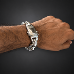 A close-up of a person's wrist wearing a chunky silver-toned chain bracelet with a rectangular clasp, set against a dark gray gradient background.