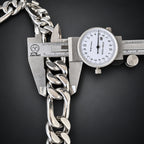 A silver-toned chunky curb chain link bracelet being measured with a mechanical caliper against a dark background.
