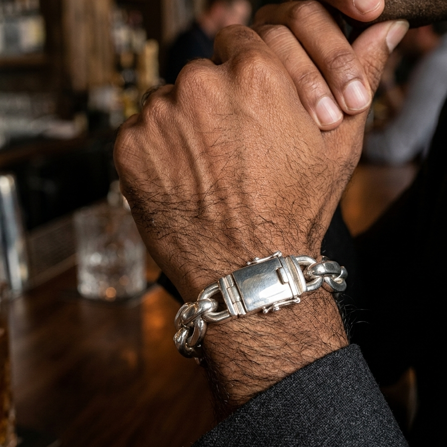 A close-up of a man's wrist wearing a chunky silver chain bracelet with a rectangular clasp, set against a blurred bar background.